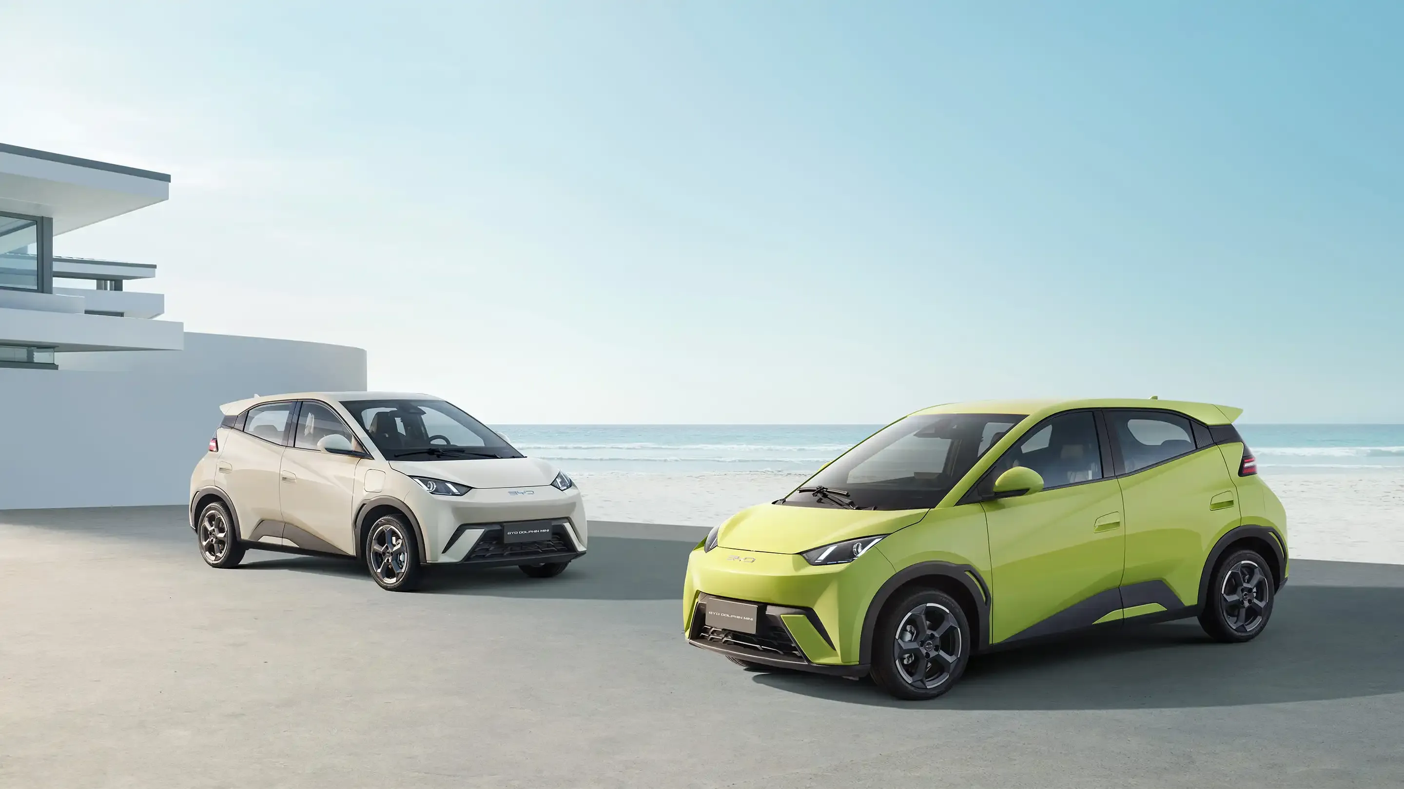 BYD Dolphin Mini verde estacionado em frente à praia, ao lado da versão branca